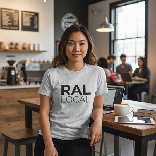 Young woman wearing RAL Local t-shirt in a cozy Raleigh coffee shop, seated at a wood table with a laptop and cappuccino