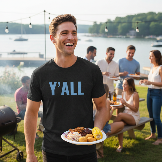 Man wearing Y'ALL shirt at outdoor cookout by the lake, smiling and holding a plate of food